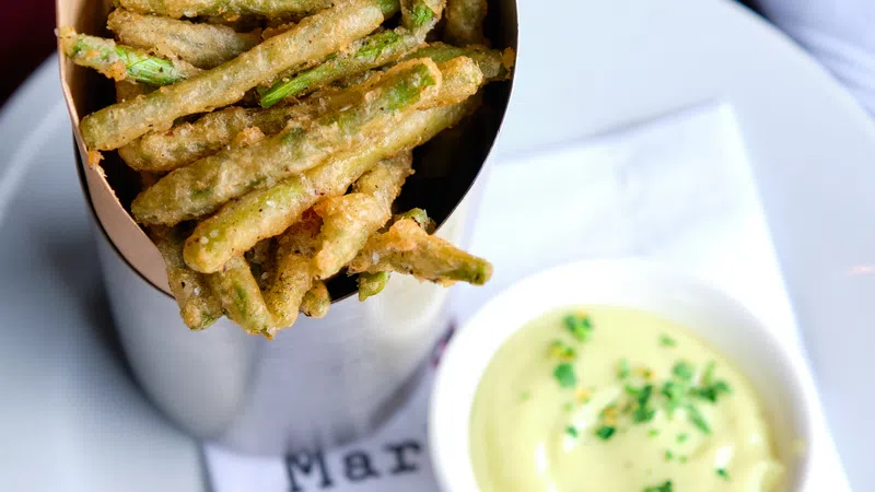 Crispy fried green beans served in a metal container with dipping sauce on a white plate and napkin.