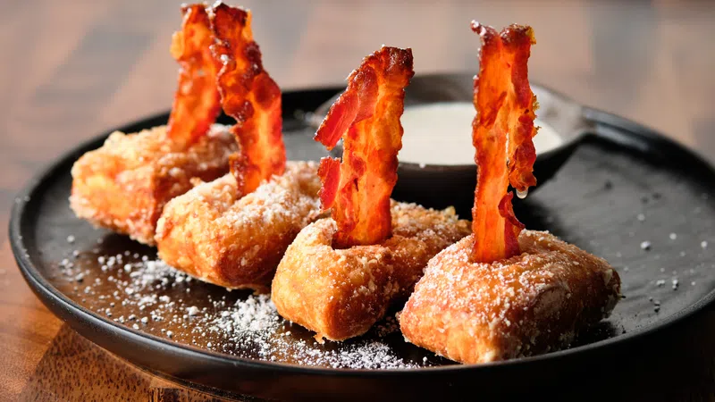 Fried dough topped with crispy bacon strips dusted with powdered sugar served on a black plate with dipping sauce.