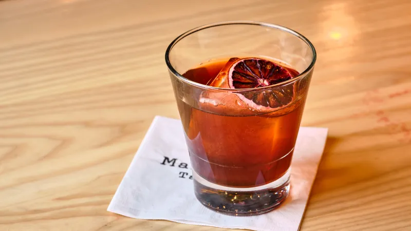 Whiskey cocktail with large ice cube and blood orange slice on a napkin on wooden table.