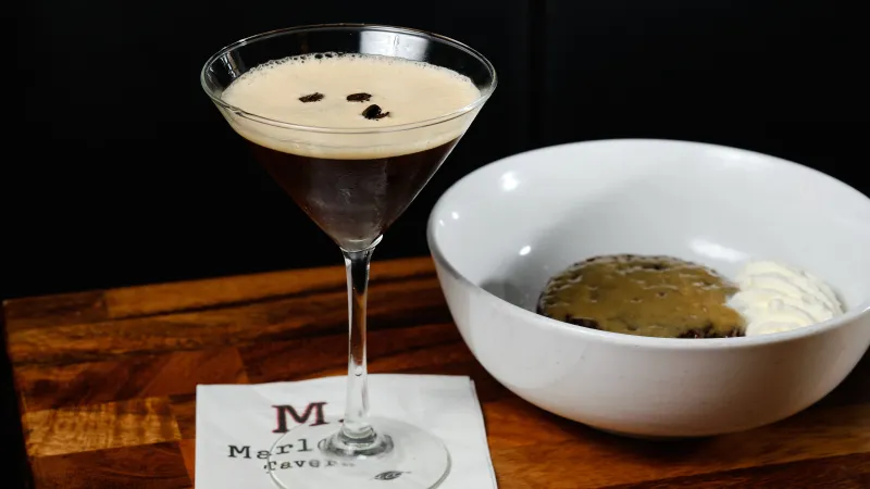 Espresso martini in a cocktail glass next to a cookie with whipped cream in a white bowl on wooden table.