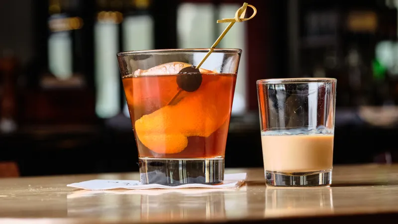Cocktail glass with an orange peel and cherry next to a small creamy drink on a wooden bar surface.