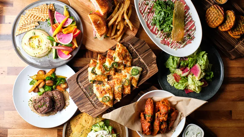 Overhead view of assorted dishes including flatbread, wings, salad, steak, sandwiches, and dips on a wooden table.