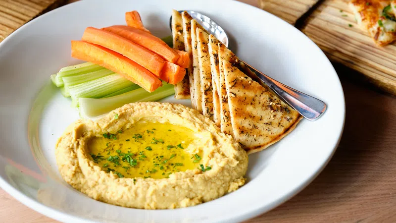 Plate of hummus garnished with olive oil and herbs served with carrot sticks, celery, and grilled pita bread.