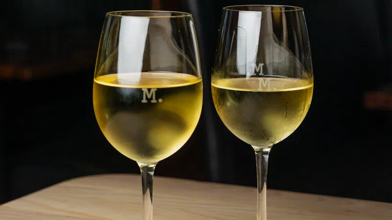 Two glasses of chilled white wine on a wooden table in a dimly lit setting with an M engraved on each glass.