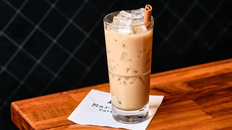 Tall glass of iced coffee with milk and cinnamon stick on wooden table with napkin