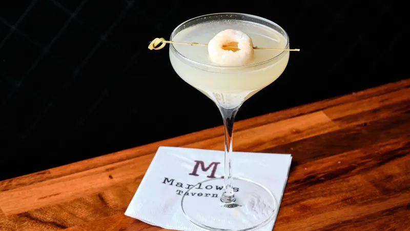 Cocktail in a coupe glass garnished with a lychee on a skewer on a Maylow's Tavern napkin on a wooden table.