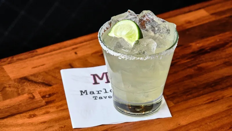 Cold margarita cocktail with lime wedge and salted rim served on a wooden bar counter on a napkin.