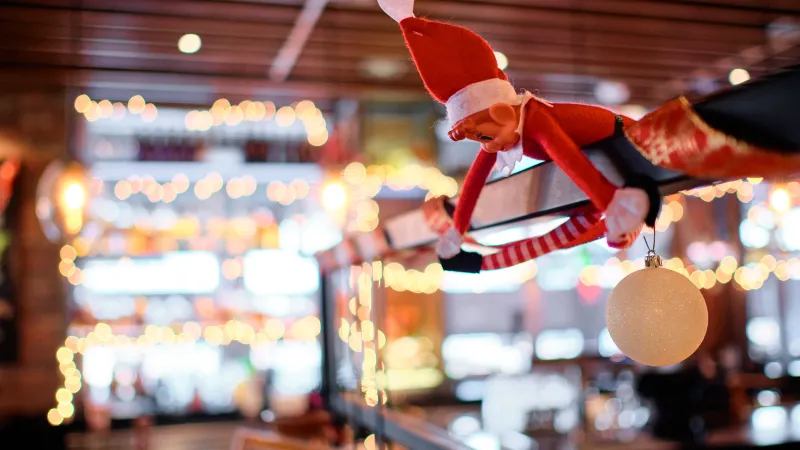 Festive elf doll hanging on a railing with Christmas lights and blurred holiday decorations in the background