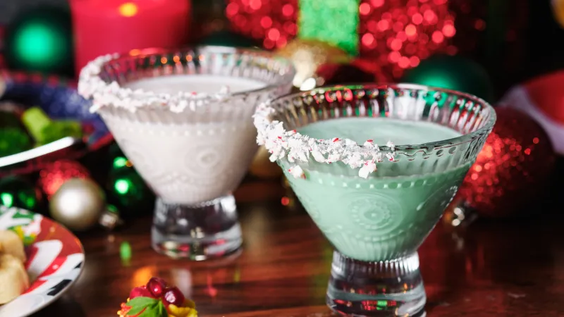 Two festive holiday cocktails in decorative glasses with crushed peppermint rims near Christmas ornaments and candles.