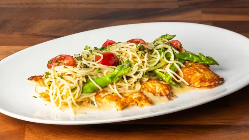 Grilled chicken breast topped with spaghetti, asparagus, and cherry tomatoes on a white plate over wooden table.