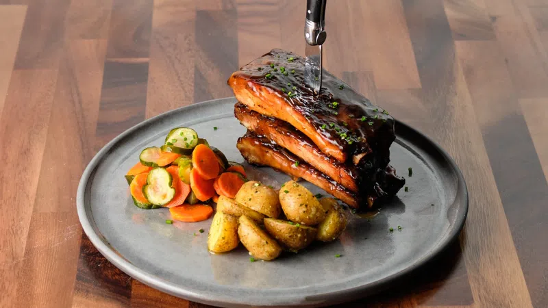 Barbecue ribs with knife inserted on plate served with roasted potatoes and mixed vegetables on wooden table.
