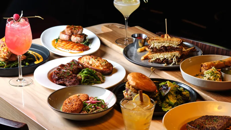 Table filled with a variety of plated gourmet dishes and colorful cocktails at a restaurant setting.