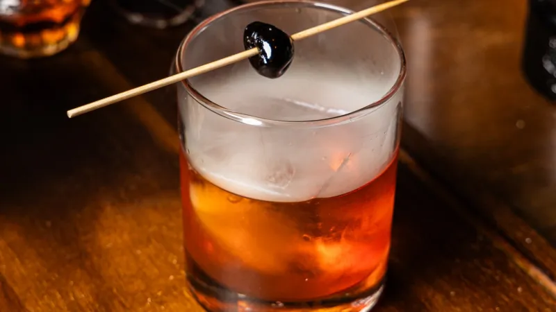 Glass of classic Old Fashioned cocktail with cherry garnish on wooden bar counter with bottles in background