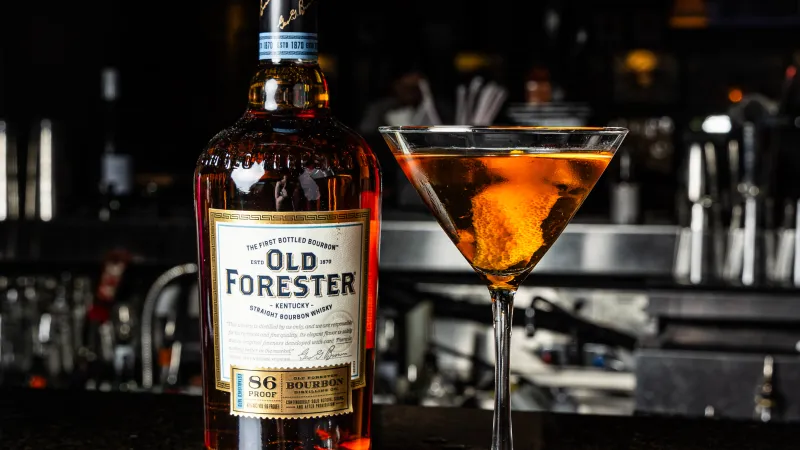 A bottle of Old Forester bourbon next to a classic cocktail in a stylish bar setting.