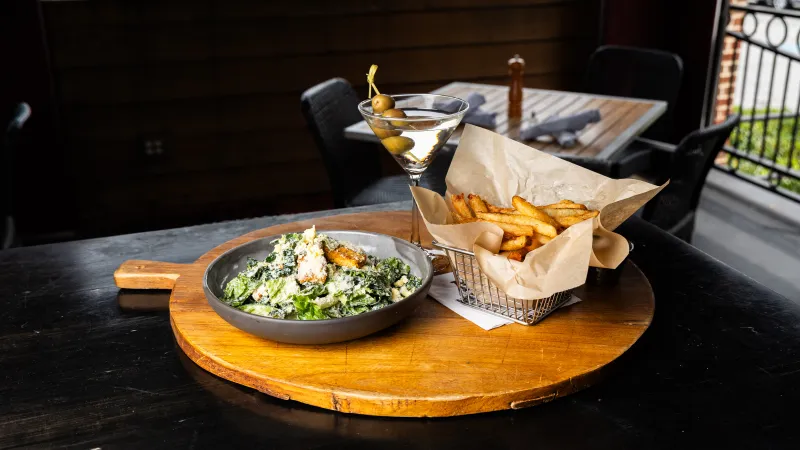 Caesar salad, basket of fries, and martini with olives served on wooden board at outdoor table