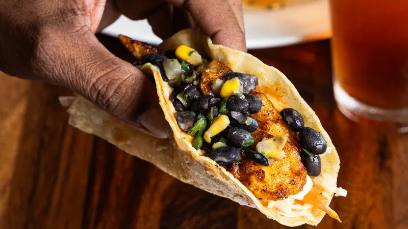 Hand holding a fish taco with black beans, corn, and vegetables over a white plate with another taco and a glass of iced tea.