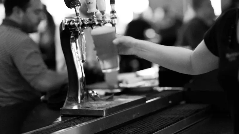 Black and white photo of a bartender pouring draft beer into a glass at a bar counter with blurred patrons.