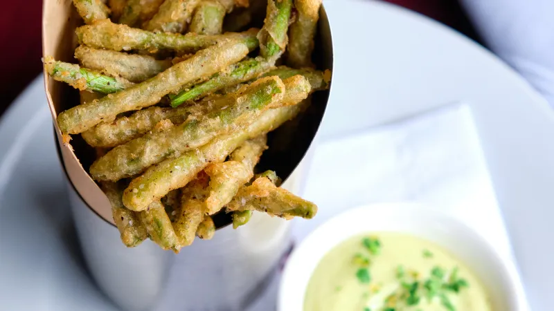 Crispy fried green bean fries served in a paper cone with a side of creamy herb dipping sauce.