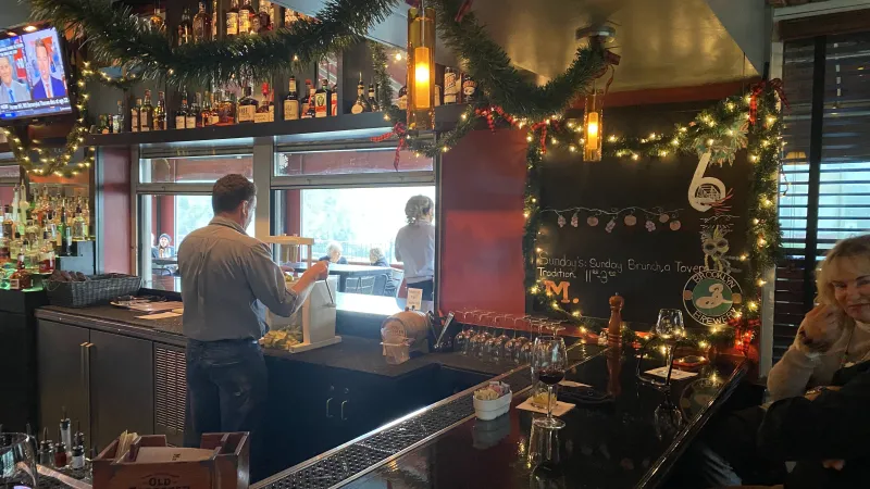 Cozy bar interior decorated with Christmas garlands, a bartender, and patrons enjoying drinks near a window.