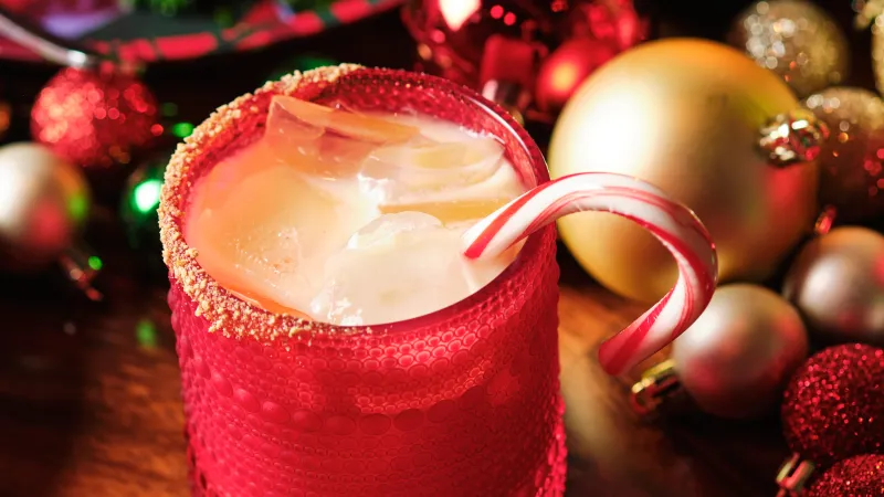 Festive holiday drink in a red glass with a candy cane, surrounded by Christmas ornaments and holiday food.