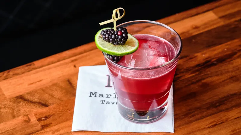 Red cocktail with ice, garnished with lime slice and blackberries on a wooden table and white napkin.