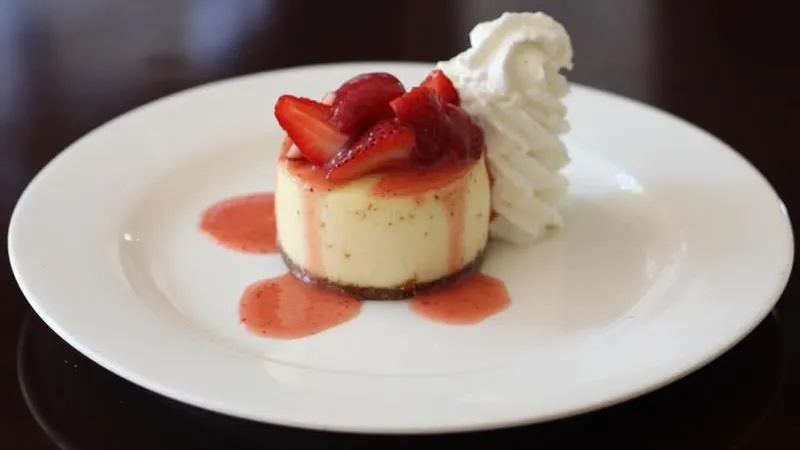 Mini cheesecake topped with fresh strawberries and red sauce served with whipped cream on a white plate