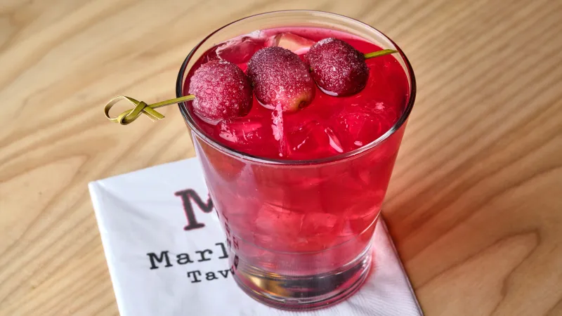 Red cocktail with ice and three cranberries on a skewer, placed on a Marly Tavern napkin on a wooden table.