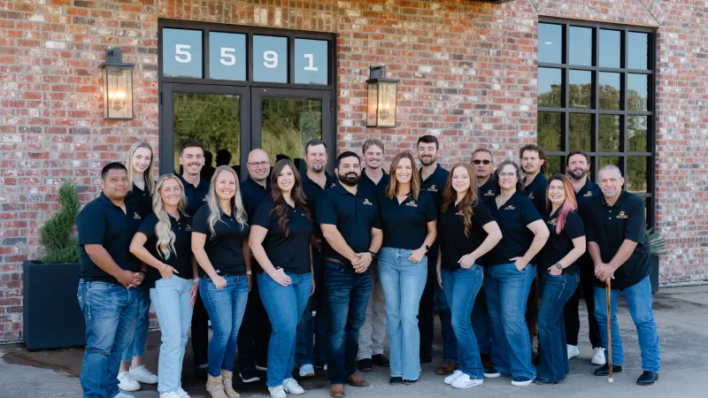 Group of 18 diverse colleagues wearing black shirts and jeans posing outside a brick building with address 5591.