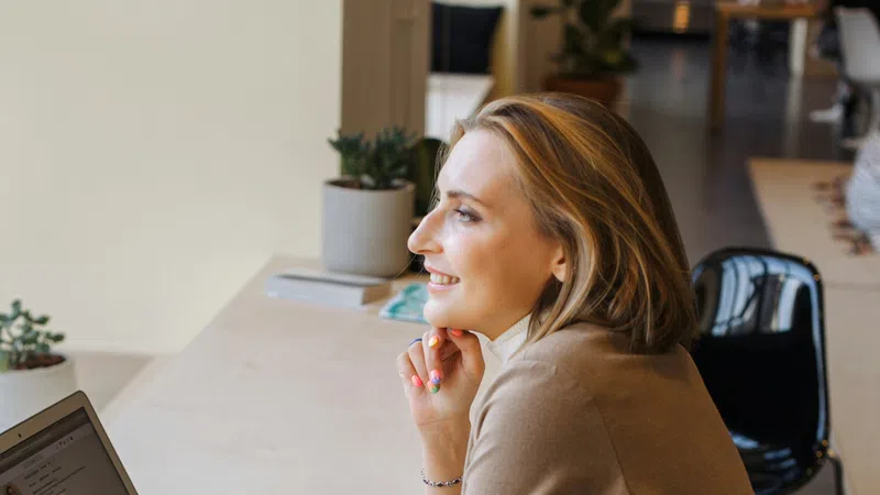 Smiling woman in beige blazer using laptop at a modern workspace with phone and plants nearby