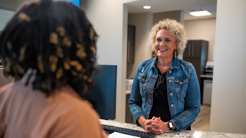 Smiling receptionist in denim jacket assisting a visitor across a marble countertop with computer and notepad.