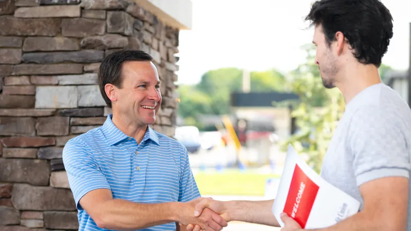 Two men shaking hands outside a building, one holding a welcome brochure, smiling and greeting.
