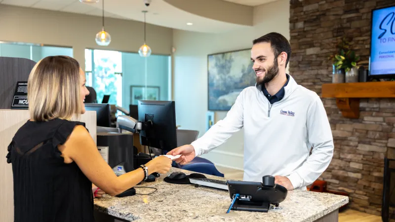 Customer handing a card to a smiling male bank teller at a modern bank counter with computers and decor.