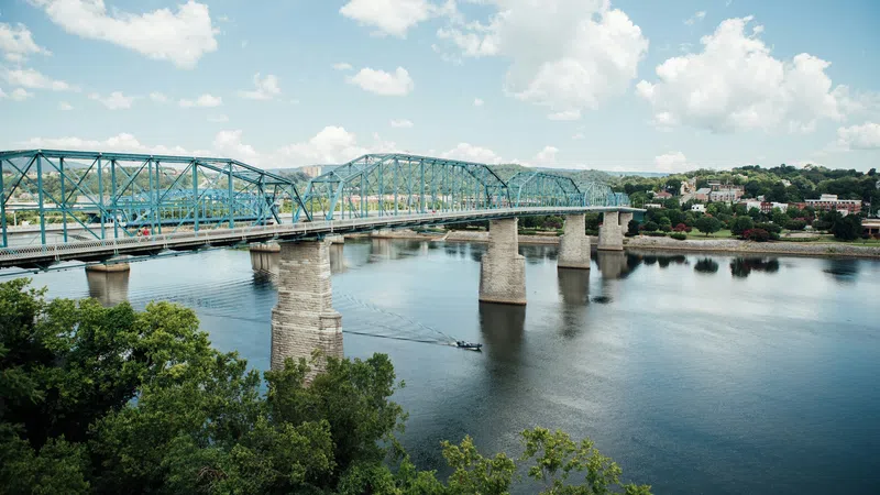Blue steel truss bridge spanning calm river with trees and cloudy sky in the background