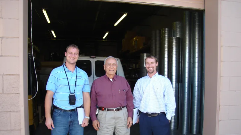 Three men standing and smiling inside a warehouse with industrial pipes and a white van in the background.