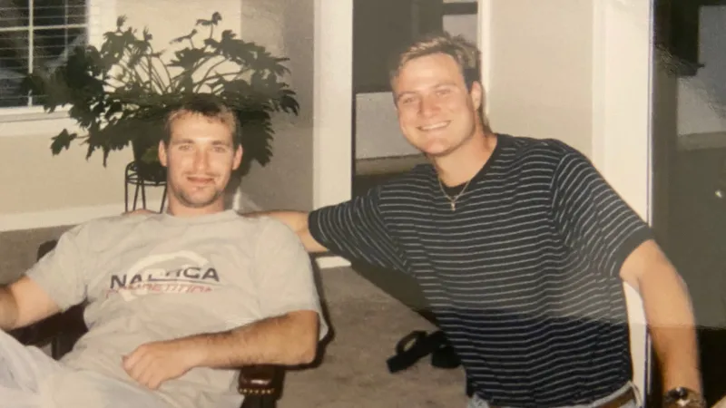 Two young men smiling indoors, one seated with a drink, the other leaning on a chair in casual clothes.