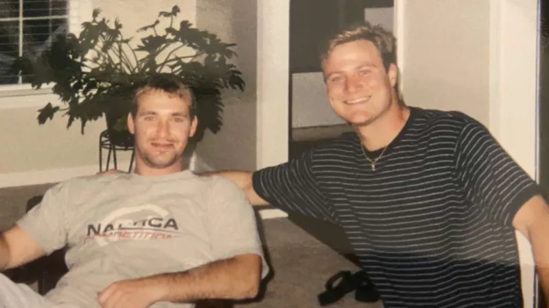 Two young men smiling indoors, one seated in a chair wearing a Nautica shirt, the other leaning forward in a striped shirt.