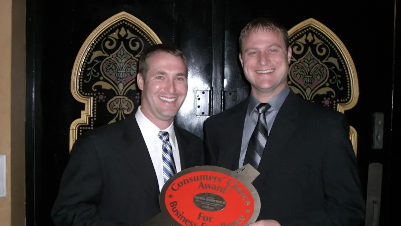 Two men in suits smiling and holding a Consumers' Choice Award for Business Excellence plaque indoors.