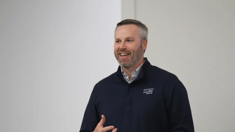 Smiling man in navy blue Infinity jacket speaking and gesturing against a plain light background
