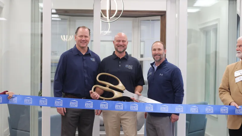 Three men in an indoor office setting conducting a ribbon-cutting ceremony with oversized gold scissors.