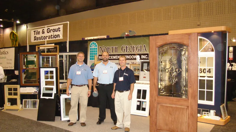 Three men standing in front of North Georgia Replacement Windows trade show booth with door and window displays.