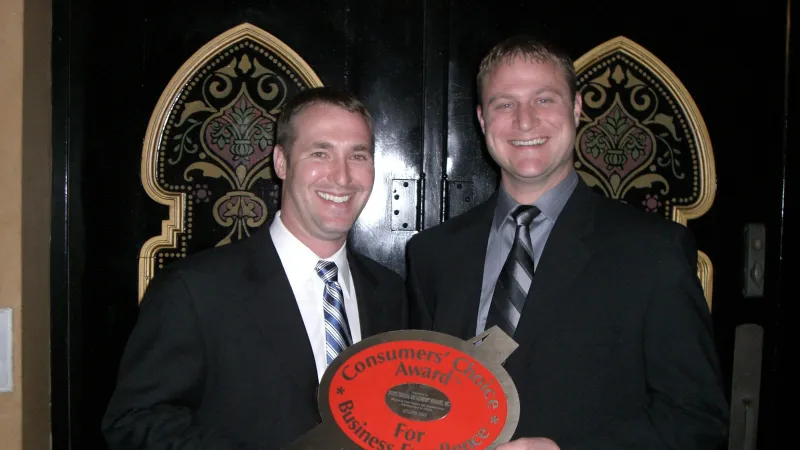 Two men in suits smiling and holding a Consumers' Choice Award for Business Excellence plaque indoors.