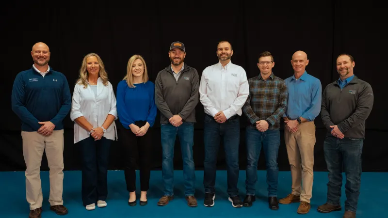 Group of eight professionals standing in a row against a black backdrop on a blue floor, dressed in business casual attire.