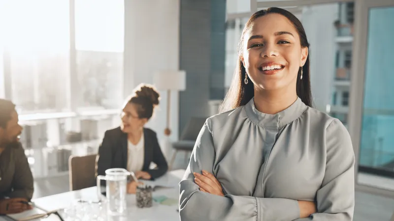 Confident young woman standing with crossed arms in modern office with colleagues talking in background