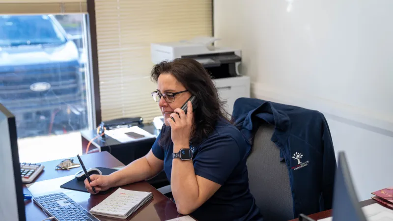 Woman in glasses talking on phone and working at desk with keyboard, notebook, and office printer in background