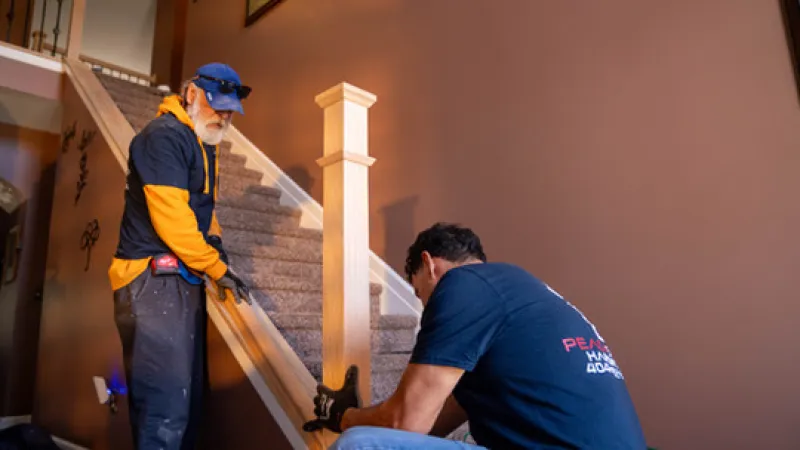 Two workers installing a wooden handrail on an indoor staircase in a home with brown walls.