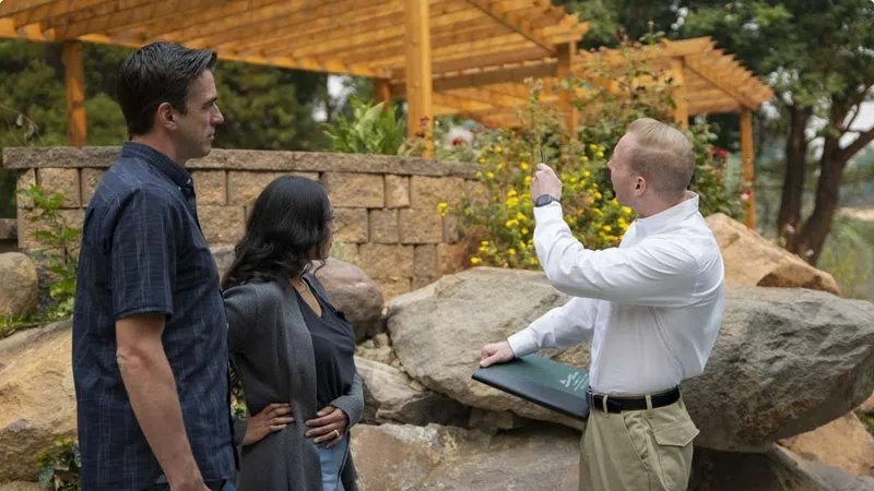 Real estate agent showing outdoor garden space to interested couple near stone wall and pergola.