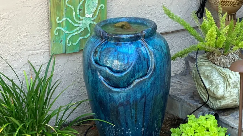Blue pottery water fountain surrounded by green plants and garden decor on a stone patio.