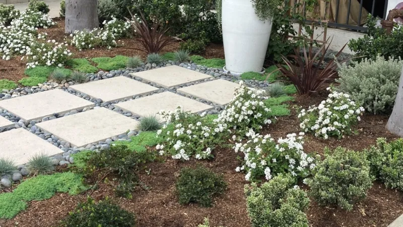 Modern garden with square stone pavers, white flowering plants, shrubs, and large white planter near palm trees.