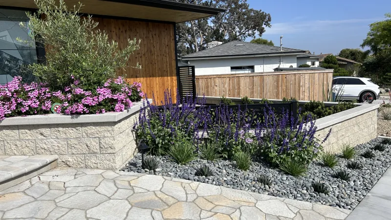 Modern house exterior with stone patio, raised flower beds with purple and pink flowers, and gravel landscaping.