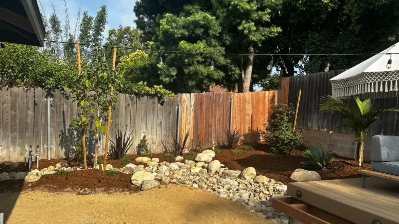 Sunlit backyard with gravel pathways, rock garden, wooden deck, outdoor seating, and greenery along a wooden fence.